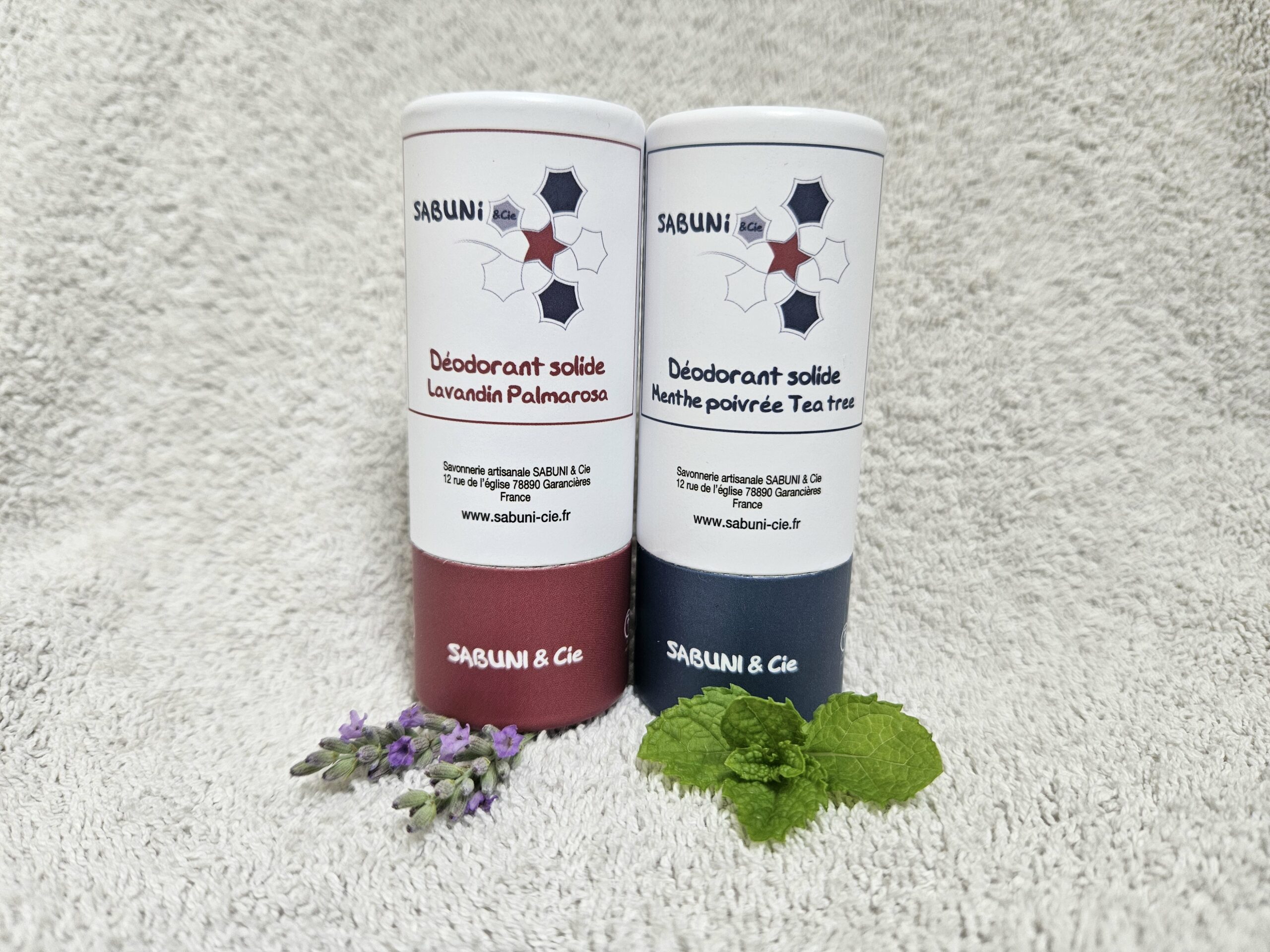 Photo des 2 déodorants posés sur une serviette. Celui parfumé au lavandin et palmarosa a un socle bordeaux. Celui parfumé à la menthe poivrée et tea tree a un socle bleu foncé.