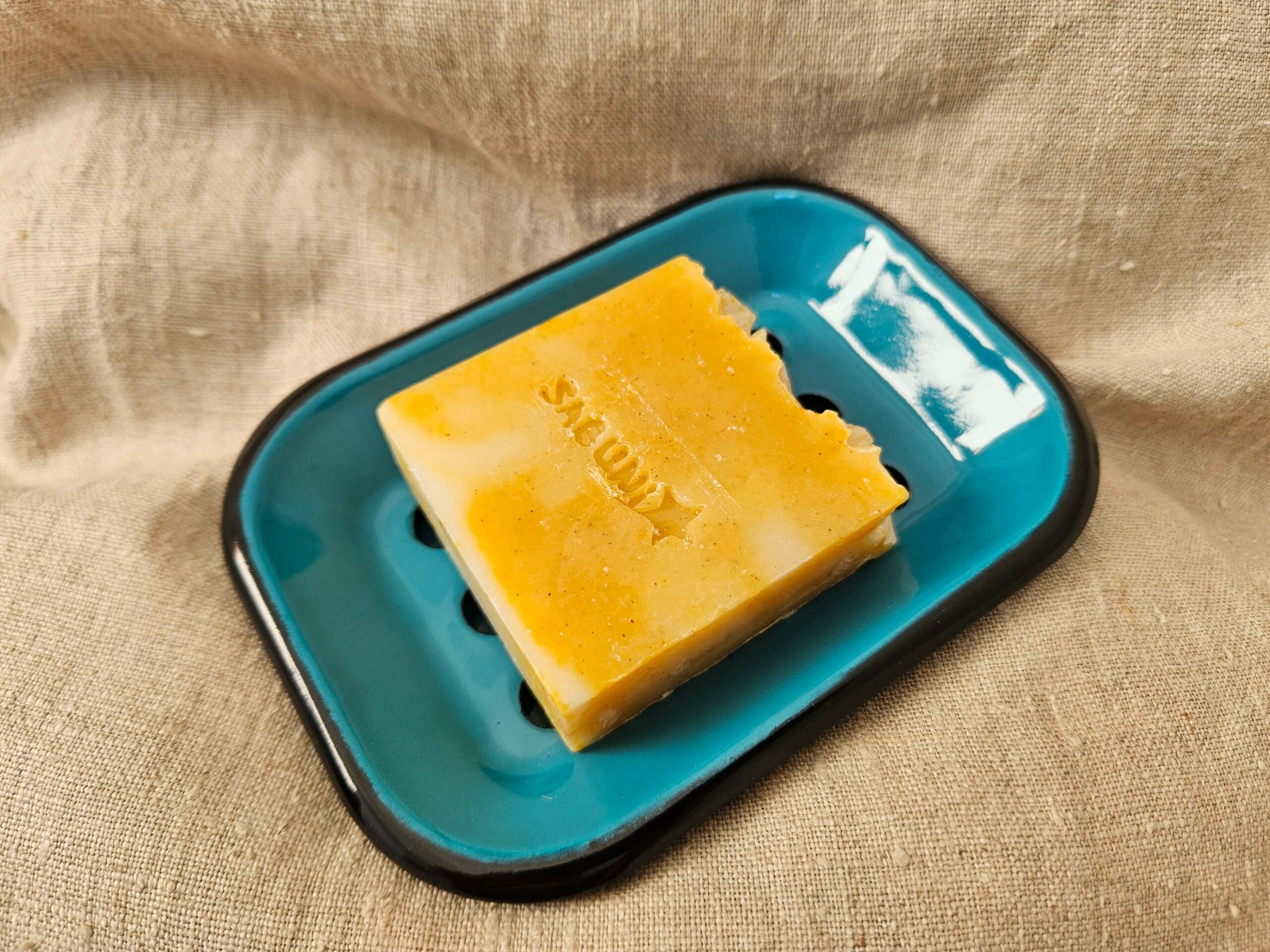 Accessoire zéro déchet de la savonnerie artisanale Sabuni & Cie. Porte-savon en fer émaillé fabriqué en Europe. Porte-savon en fer émaillé sur fond en fibre naturelle.