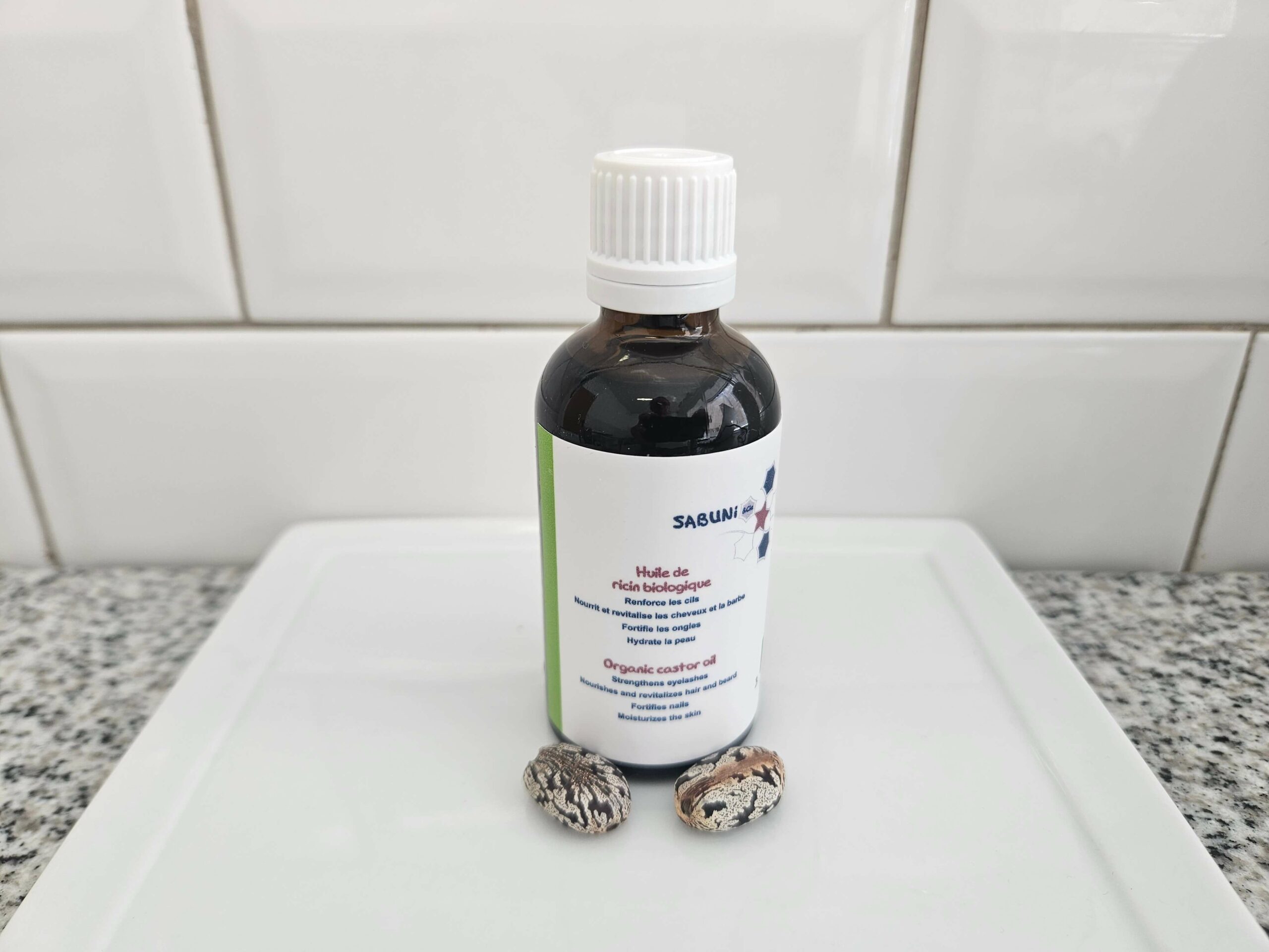 Huile de Ricin biologique, reconditionnée par la savonnerie artisanale Sabuni & Cie, dans un flacon en verre opaque, posé devant du carrelage style métro.